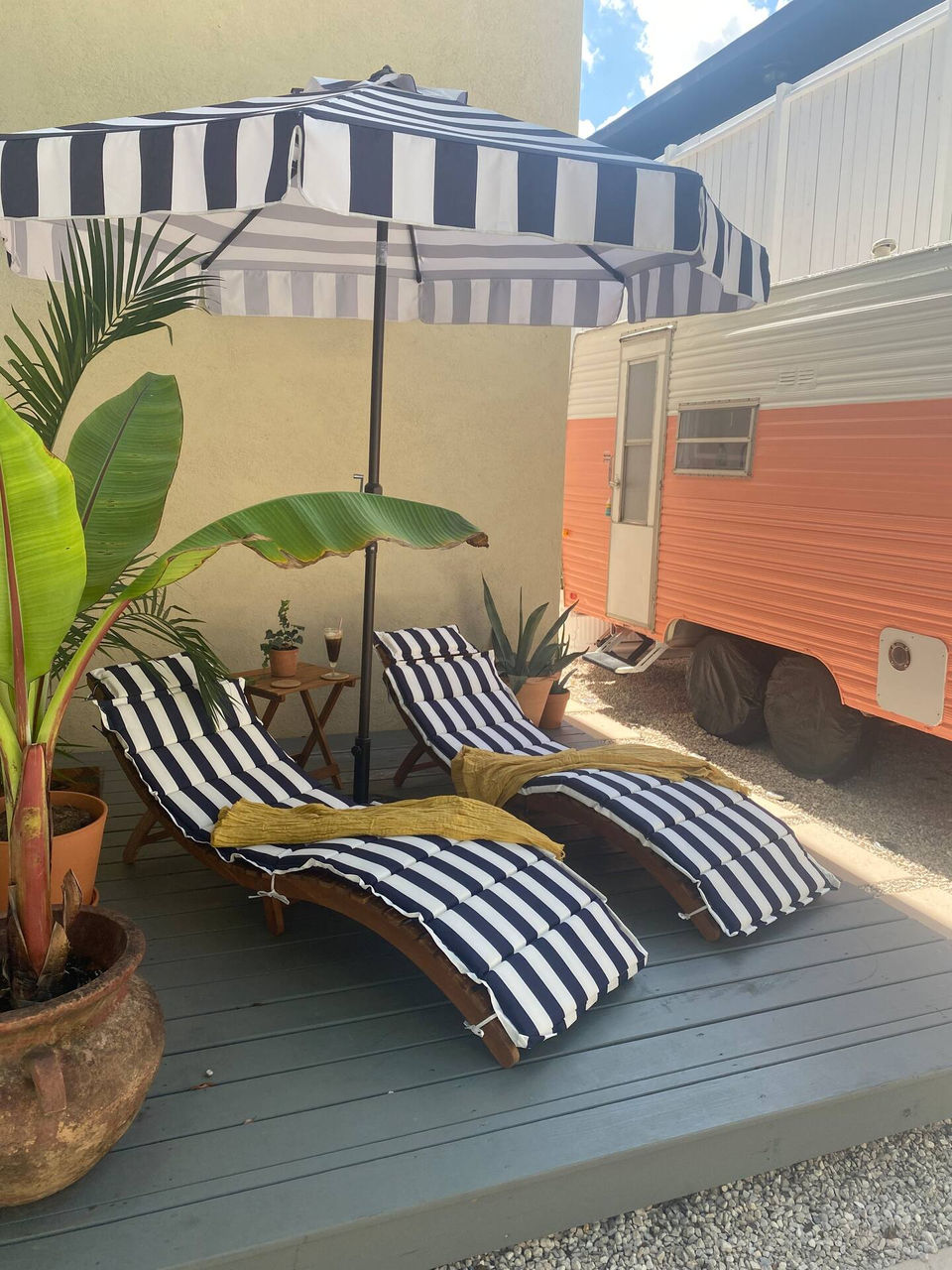Two lounge chairs under a striped umbrella nestled together on a deck adjacent to Poppy, the Vintage Glamper/RV.