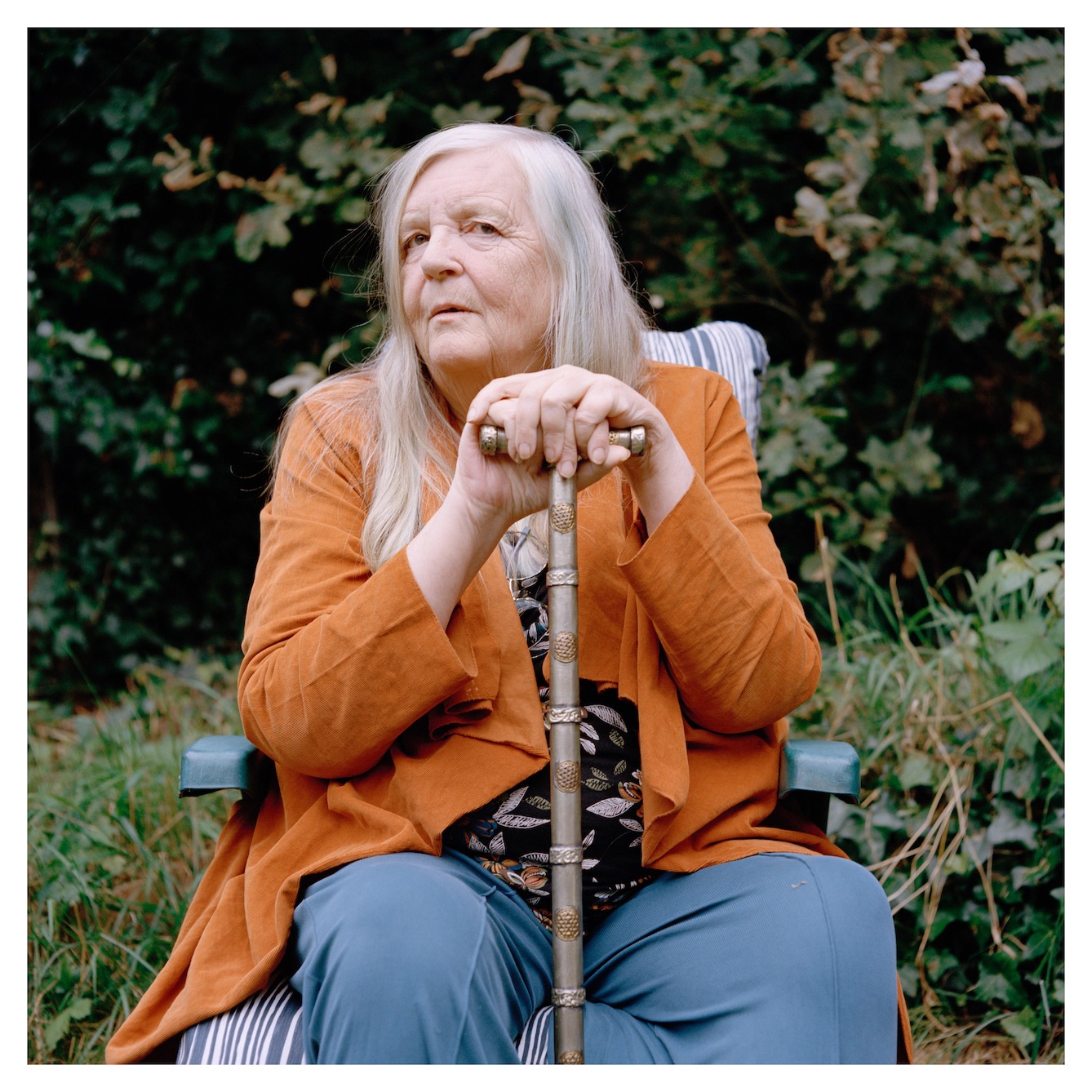 Woman seated with grey, long hair, orange sweater, blue jeans, multi-colored shirt, and cane. Landscape is in the background.