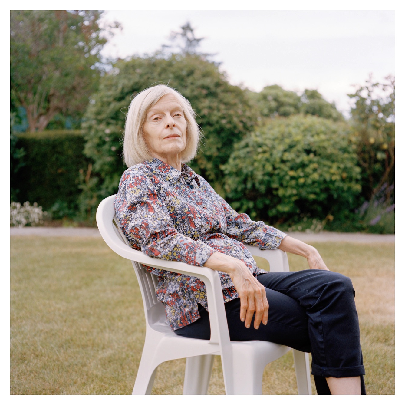 Woman looking at camera with blonde bob, flowered long sleeve shirt, and black pants. In a white chair with landscaping in the background.