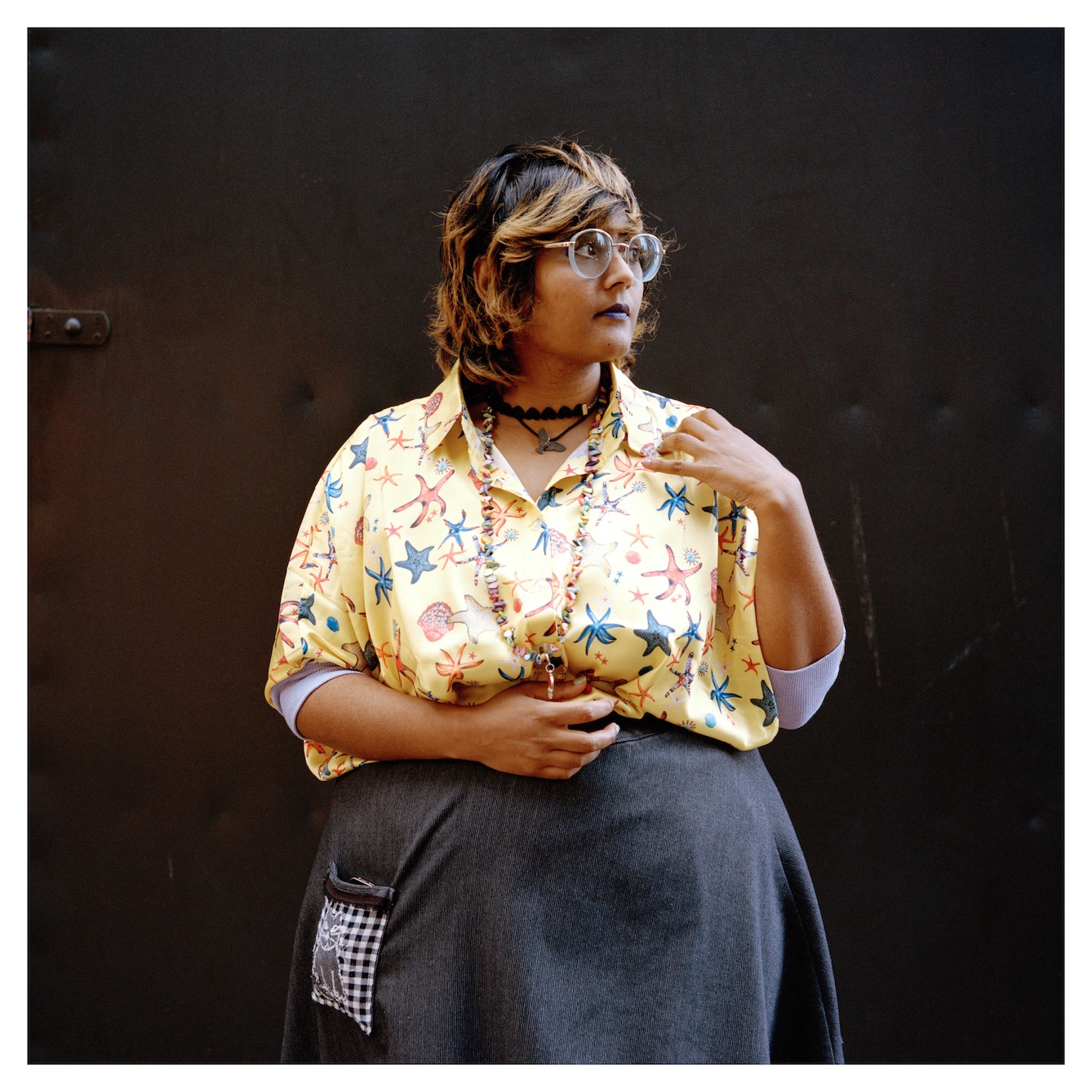 Adult with golden hair looking to the right. Individual has large glasses, multiple necklaces, yellow shirt with stars, and long skirt.