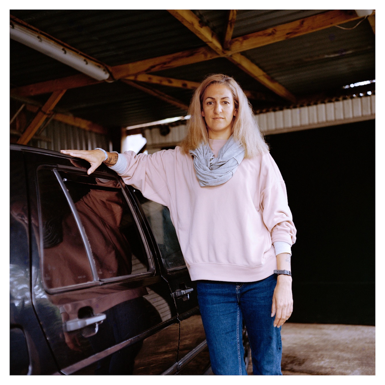 Adult with long blonde hair, pink shirt, purple scarf, and blue jeans, leaning agains black car.