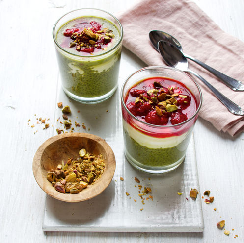 Zwei Gläser mit geschichtetem Matcha-Chia-Pudding, weißem Joghurt und leuchtend rotem Himbeer-Püree, bestreut mit gehackten Pistazien auf einem hellen Holztisch.