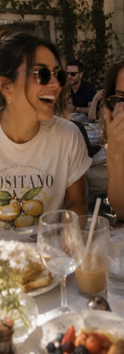 Woman laughing with a friend at an outdoor European café while wearing a white Positano lemon graphic tee styled for summer lunch and vacation outfits.