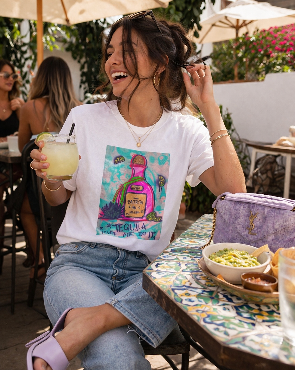 Woman sitting at an outdoor café holding a cocktail while wearing a “Tequila Made Me Do It” graphic tee, styled with denim for a relaxed girls day outfit