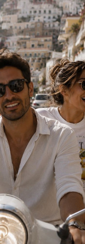 Couple riding a scooter along the Amalfi Coast with woman wearing a white Positano lemon graphic tee, sunglasses, and vacation-ready summer styling.