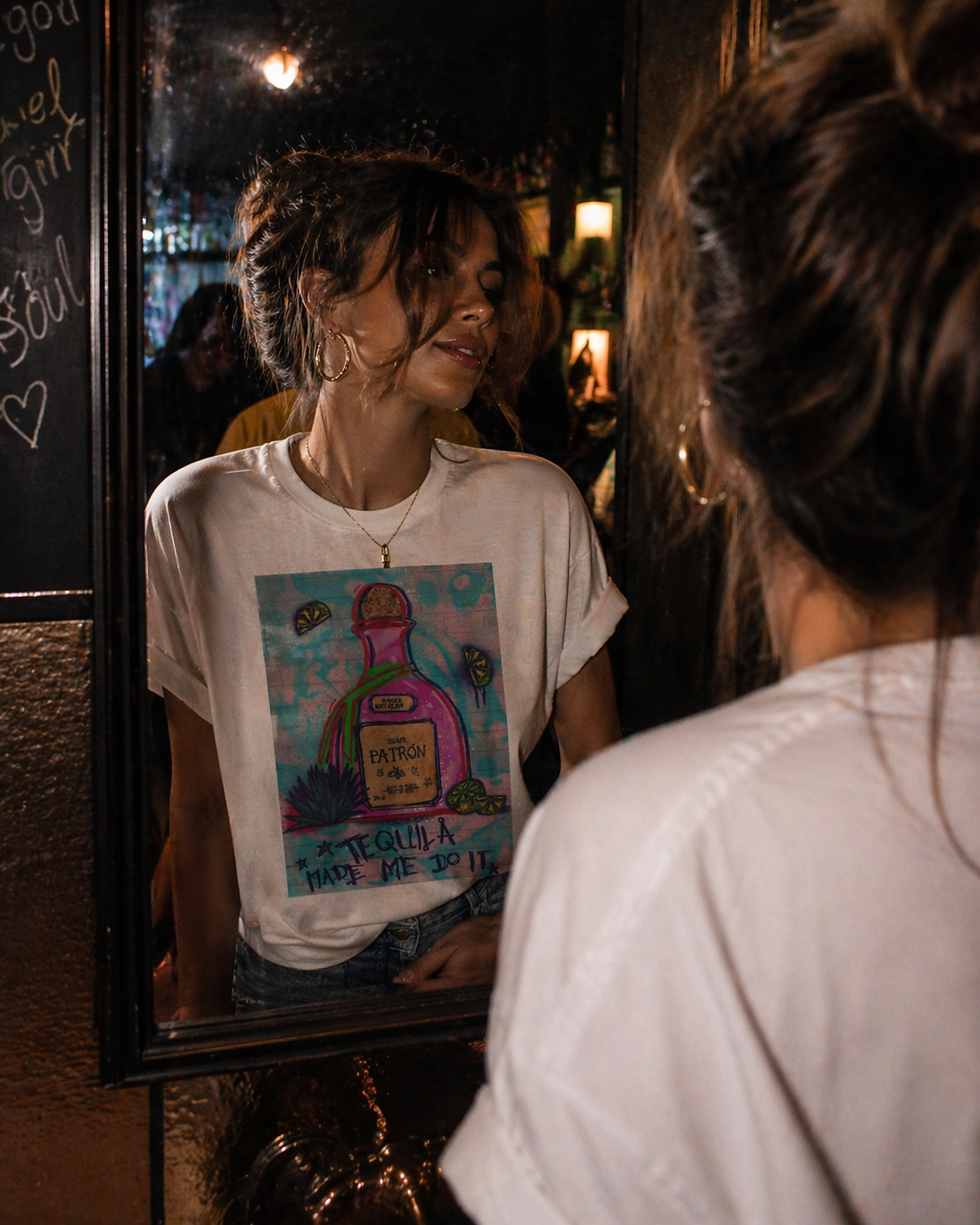 Woman in a dimly lit bar mirror wearing a “Tequila Made Me Do It” graphic tee, messy hair and glowing skin, capturing a confident, carefree night out moment