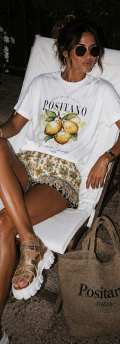 Woman wearing a white Positano lemon graphic tee with patterned shorts, gold sandals, sunglasses, and a woven tote while lounging on a sunny Italian patio with espresso cups nearby.