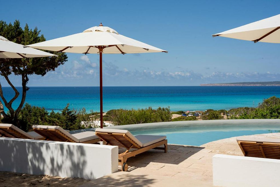Sun loungers under white umbrellas overlook a serene pool and vibrant blue ocean in Formentera, Spain, with a clear sky and lush greenery in the background, at Dunas de Formentera Luxury Hotel.