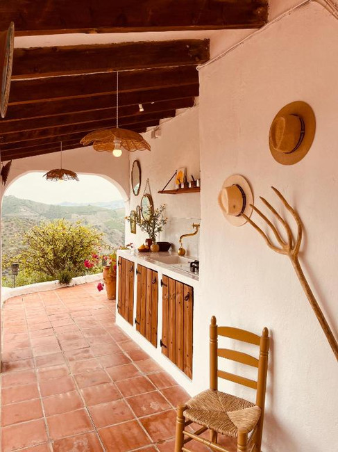 Rustic outdoor kitchen with wooden cabinets, a chair, straw hats, and a scenic view of hills and greenery beyond at Casita Comares Adults Only Hotel in Costa del Sol