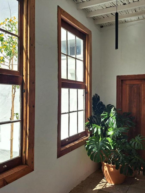Sunlit corridor with large wooden-framed windows, revealing a quaint courtyard filled with vibrant flowers. A potted plant sits beside a rustic wooden door at Casa de las Flores adults only hotel in Lanzarote