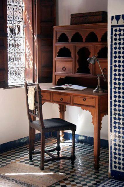 A rustic wooden desk and chair in a bright room, featuring intricate tiled flooring and sunlight streaming through patterned windows at Riad Seffarine in Fez.