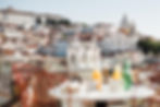 Rooftop view of Lisbon's Alfama district with red-tiled roofs, historic white buildings, and blurred bottles on a sunny terrace.