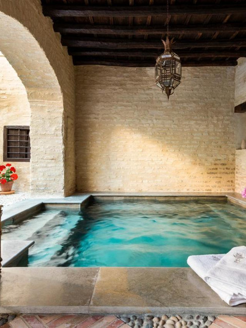 A serene indoor pool area featuring a lantern, fresh flowers, and potted geraniums against textured stone walls at Palacio Bucarelli, a hotel with pool in Seville, Spain