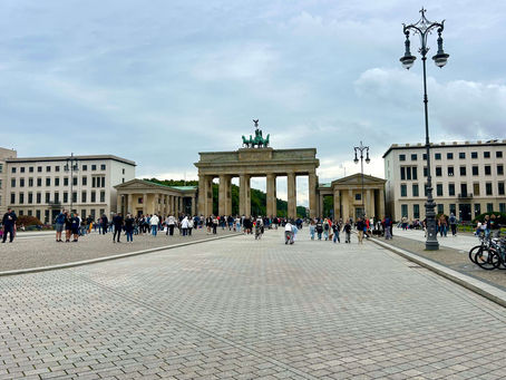People walk near the Brandenburg Gate in Berlin, under a cloudy sky. The monument is flanked by historic buildings and street lamps.