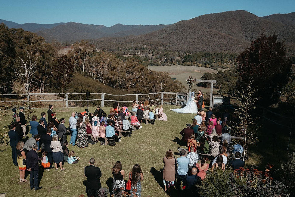 Outdoor wedding ceremony with a bride and groom under a timber arbour. Guests seated on The Lawn with mountain views in the background, sunny day.