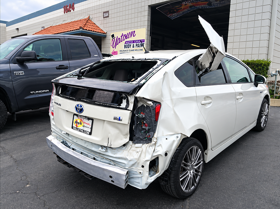White Toyota Prius with extensive rear collision damage, missing rear glass, and exposed rear bumper structure outside an auto body shop.