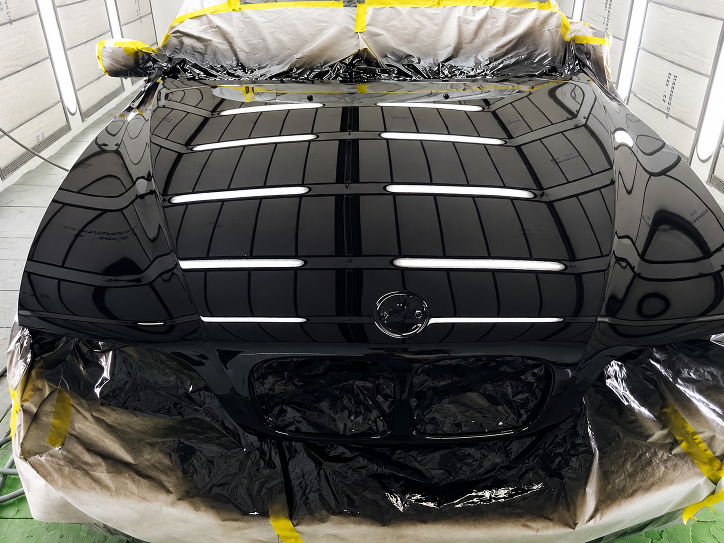 Glossy black vehicle hood and grille inside an illuminated automotive paint booth with surrounding surfaces masked.