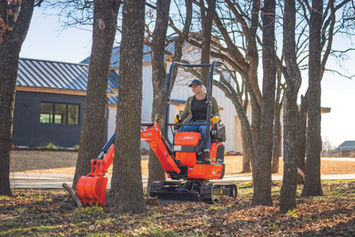 Kubota U10-5 mini excavator operating between trees with front blade and bucket visible.
