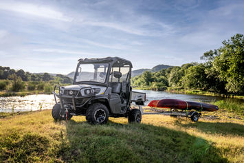 Kubota RTV-XG850 Sidekick — side outdoor view towing kayaks near river showing Stone Gray utility vehicle, trailer hitch use, roof, windshield, and ATV tires