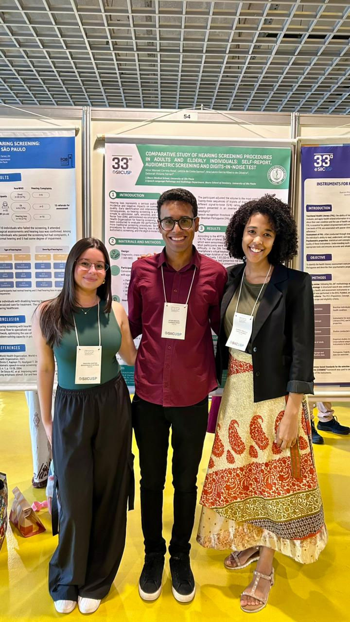 Foto de três pesquisadores sorrindo em frente aos pôsteres de seus trabalhos apresentados no SIICUSP. À direita, Isadora, mulher morena de cabelos longos e lisos, usa óculos, regata verde e calça preta. Ao centro, Vitor, homem moreno de óculos, veste camisa social vermelho-escura e calça preta. À esquerda, Queren, mulher morena de cabelo encaracolado na altura dos ombros, usa blazer escuro e saia florida. Ao fundo, aparecem os pôsteres científicos fixados em painéis.