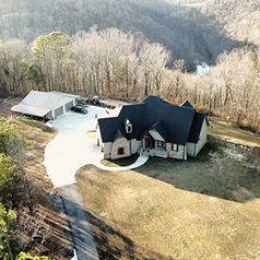 Aerial view of a secluded house with a dark roof, surrounded by leafless trees and a green forest on one side, set in a hilly landscape.