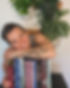 Image of a woman tilting her head and smiling while leaning on a selection of books. She has a tattoo on her forearm of leaves growing out of a book and there is a blurred out plant in the background