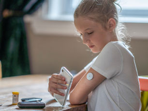 Young girl with a white shirt uses smartphone at a wooden table. Medical sensor on arm. Vial and device nearby. Bright, calm setting.