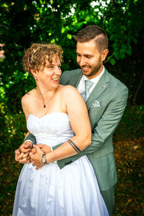 Fröhliches Hochzeitsfoto eines Brautpaars, bei dem der Bräutigam seine Braut von hinten umarmt. Beide halten sich an den Händen und lachen vor Freude. Die emotionale, natürliche Hochzeitsaufnahme zeigt Liebe, Verbundenheit und Glück.