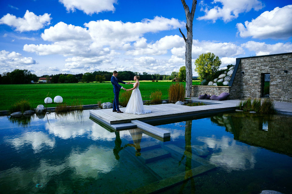 Braut und Bräutigam stehen sich auf einem Steg über einem See in den Lenderstuben Balzhausen gegenüber, umgeben von idyllischer Landschaft. Der Himmel spiegelt sich im Wasser und verstärkt die besondere Atmosphäre des First Looks.