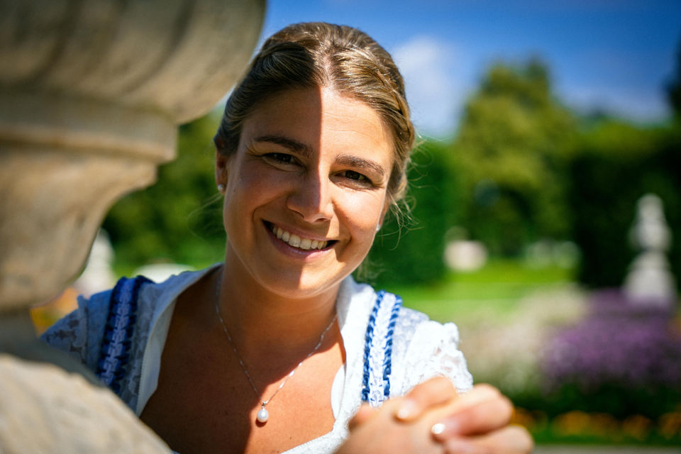Spontanes Hochzeitsfoto einer strahlenden Braut im Dirndl im Schloss Schleißheim bei München. Ihr Lächeln spiegelt pure Freude wider, während sie die Hand ihres Partners hält. Das Licht- und Schattenspiel auf ihrem Gesicht verleiht dem Moment Tiefe, während der Schlossgarten eine idyllische Kulisse bietet.
