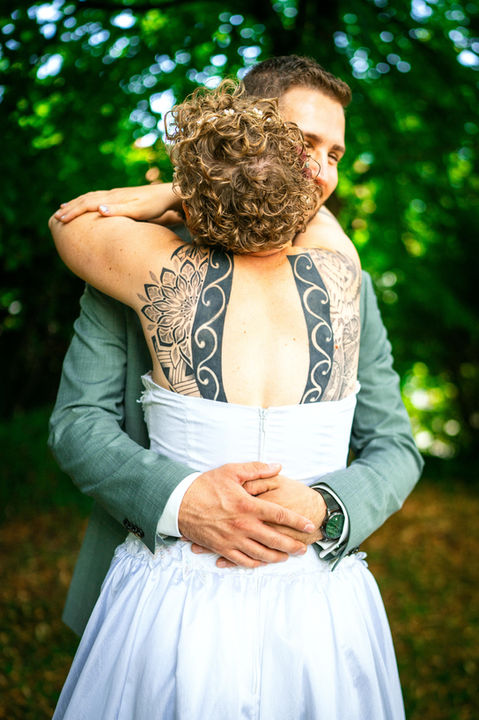 Fröhliches Hochzeitsfoto eines glücklichen Brautpaars, das sich frontal umarmt und die Hände hinter dem Rücken verschränkt. Die seitliche Aufnahme betont die innige Verbindung und das strahlende Glück des Paares in einem zeitlosen Hochzeitsmoment.