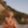 Woman with long wet light brown hair looking blankly into the distance. Striped towel wrapped around shoulders. Standing on a beach with a grassy sand dune in the back. Appears to be at dusk.