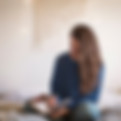 Person sitting on their bed, cross legged with a book open in their lap. They have long brown wavey hair and are wearing jeans and a dark blue jumper. Their head is tilted down and they are writing in their journal. The wall behind them is an off white colour with a wall hanging that is light brown.