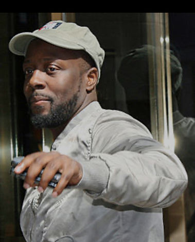 Wyclef Jean wearing a cap and jacket looks at something outside.