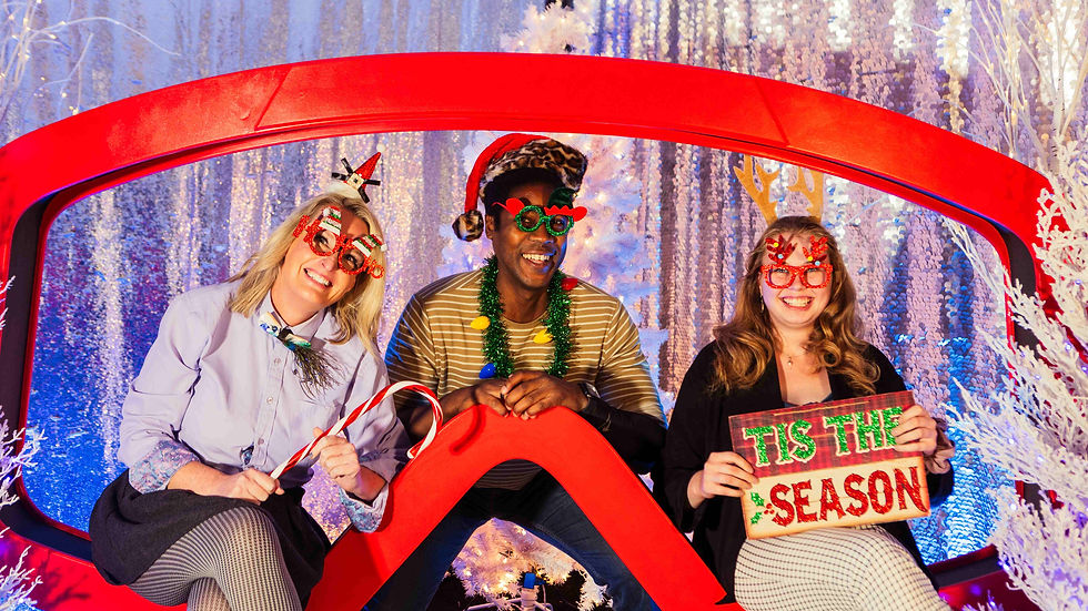 An Oversizes Ski Goggle Photo Booth, with two women and one man sitting in the center holding festive holiday signs and wearing festive holiday props taking a photo.
