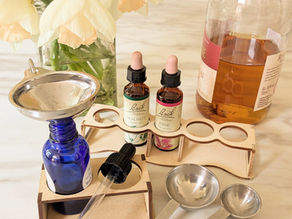 Blue bottle with metal funnel, Bach Walnut and Pine flower remedies, a bottle of brandy, measuring spoons, and daffodils in vase on a marble counter.