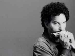 Man with curly black hair, holding harmonica and microphone to mouth.
