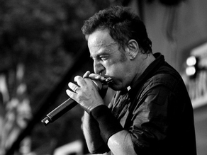 Greyscale image of man playing harmonica into microphone. He has short dark hair, a black shirt and black wristband on.