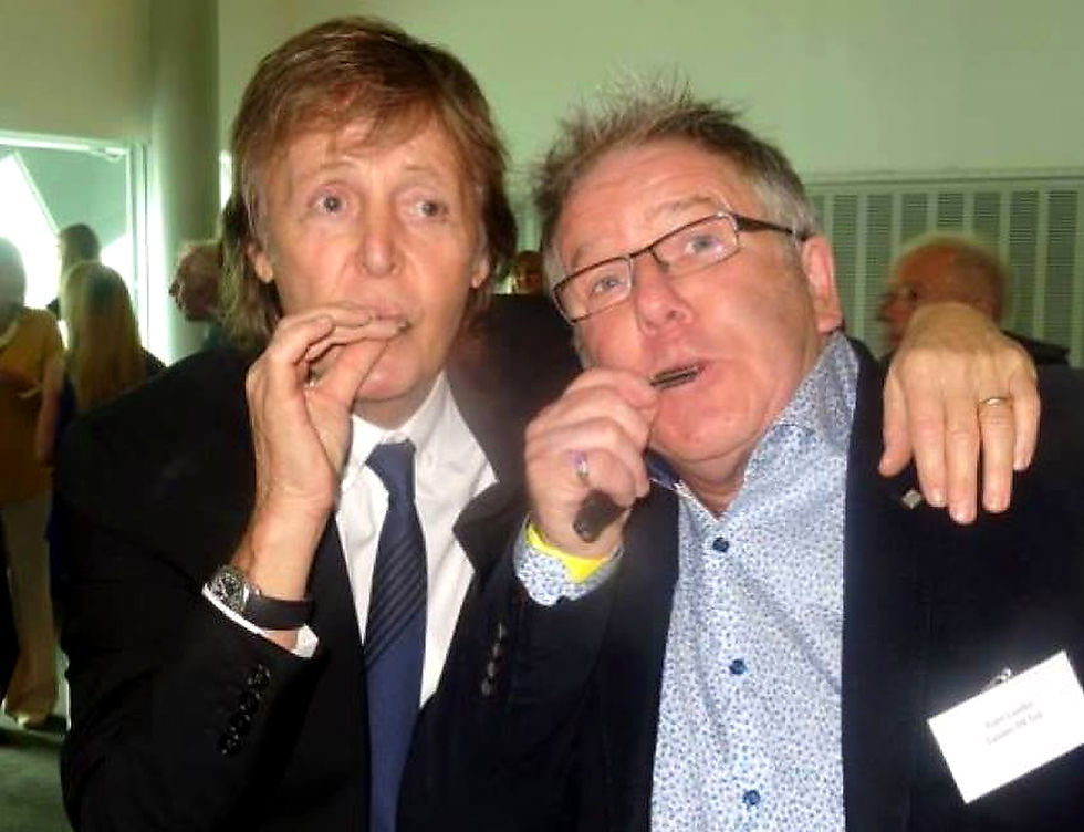 Two men in suits, playing miniature harmonicas. One man has his arm around the shoulder of the other. They are in a crowded room.