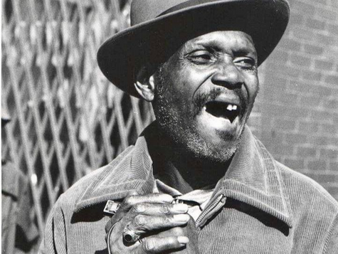 Man in hat holding harmonica in both hands, shouting or laughing in front of brick wall and fence. The photo is greyscale.