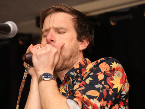 Man playing harmonica with microphone, wearing colourful shirt, eyed closed. 