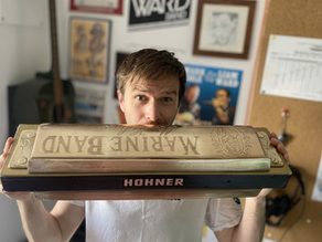 Man in a white T-shirt holds a large Hohner Marine Band harmonica toward camera in a music room, looking surprised.