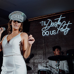 bride holding microphone with silver hat in front of til death do us party neon sign