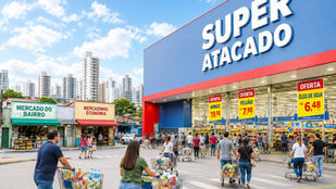 Consumidores circulando entre corredores de um grande supermercado enquanto pequenos mercados aparecem ao fundo com pouco movimento.