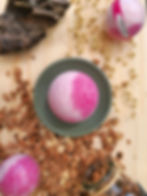 Bombe de bain framboise posée sur socle vert d'eau sur une planche en bois et des copeaux, entourée d'autres boules roses