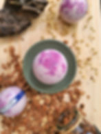 Bombe de bain Baies sauvages posée sur socle vert d'eau sur une planche en bois et des copeaux, entourée de boules violettes