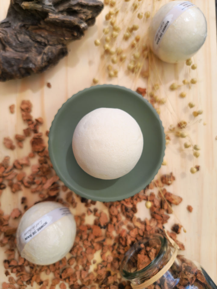 Bombe de bain Lait de chèvre posée sur socle vert d'eau sur une planche en bois et des copeaux, entourée de boules blanches