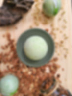 Bombe de bain kiwi posée sur socle vert d'eau sur une planche en bois et des copeaux, entourée d'autres boules vertes