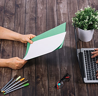 person-typing-laptop-with-his-colleague-holding-blank-white-green-blank-paper-wooden-table