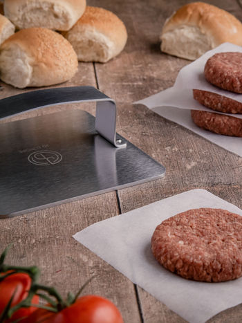 A square metal patty press produced by JD Europe styled with burger patties and buns on a worn wooden table.